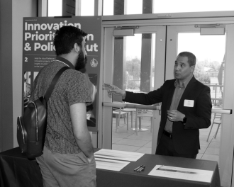 Two men have a discussion at an event booth with a sign reading “Innovation Prioritization & Policy Input.” Papers and a pen are laid out on the table in front of them.