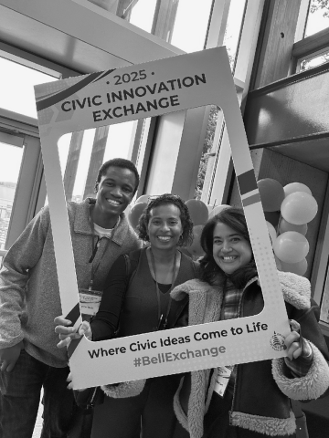 Three people smile while posing indoors with a large photo frame reading "2025 Civic Innovation Exchange".
