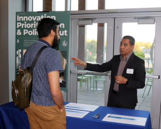Two men have a discussion at an event booth with a sign reading “Innovation Prioritization & Policy Input.” Papers and a pen are laid out on the table in front of them.