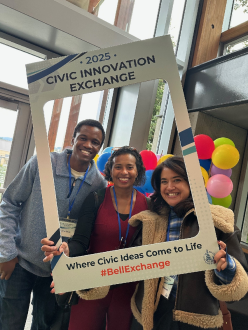 Three people smile while posing indoors with a large photo frame reading "2025 Civic Innovation Exchange".